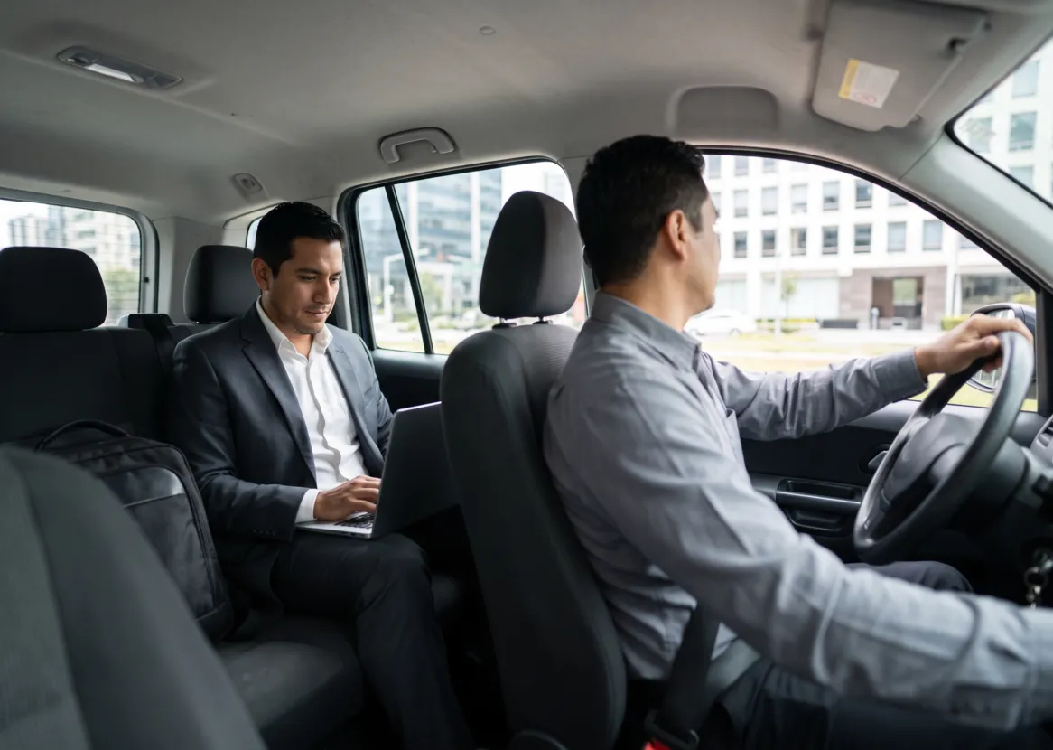 Conductor profesional al volante y ejecutivo con laptop en el asiento posterior de una camioneta (imagen referencial)