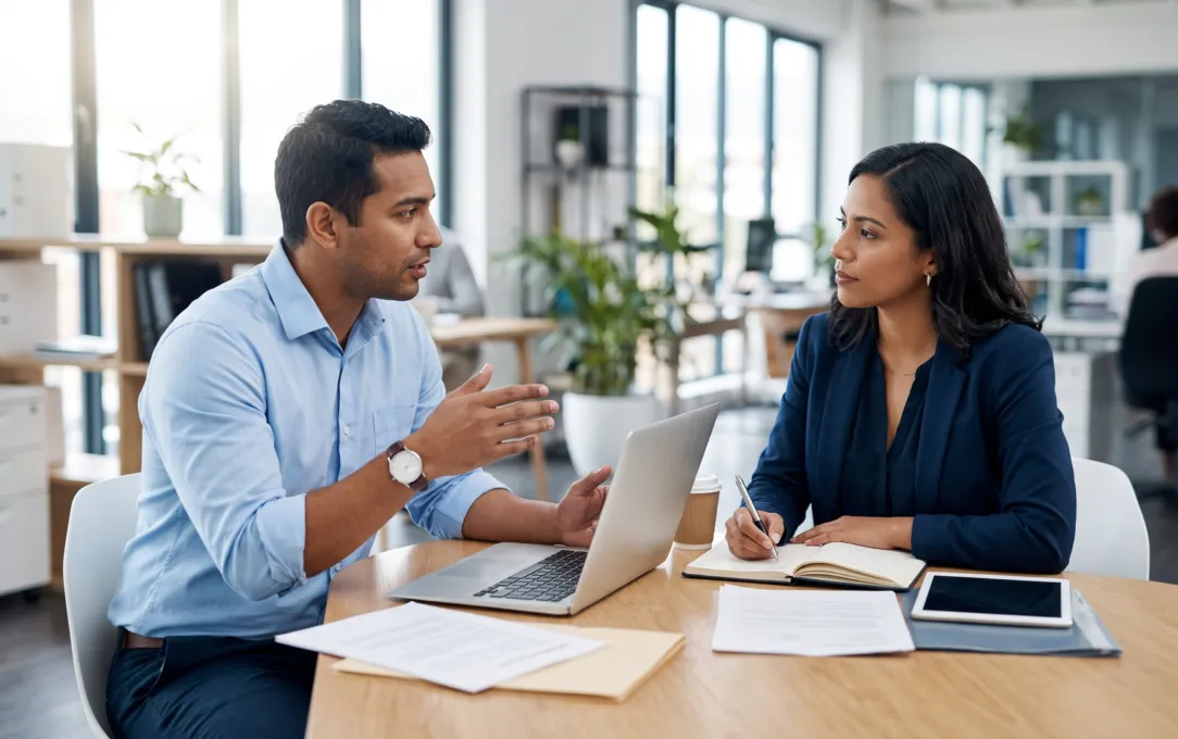 Dos profesionales conversan sobre el modelo de trabajo de la empresa, con laptop, libreta y tableta sobre la mesa (imagen referencial)